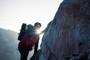 Climber with a red backpack ascending a rocky cliff, sun flashing between their hand and the rock surface at sunrise.