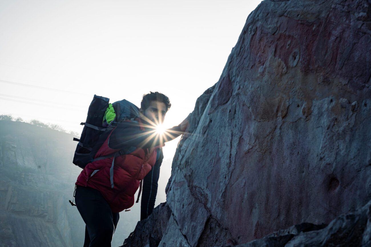 Climber with a red backpack ascending a rocky cliff, sun flashing between their hand and the rock surface at sunrise.