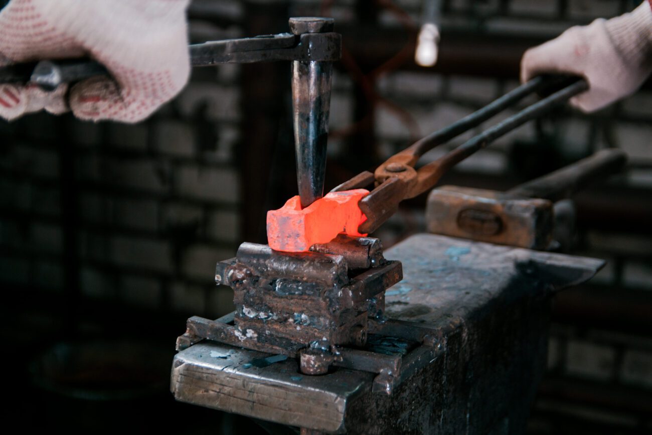 Gloved hands hold tongs to shape a glowing orange-hot metal piece on an anvil in a blacksmithing workshop