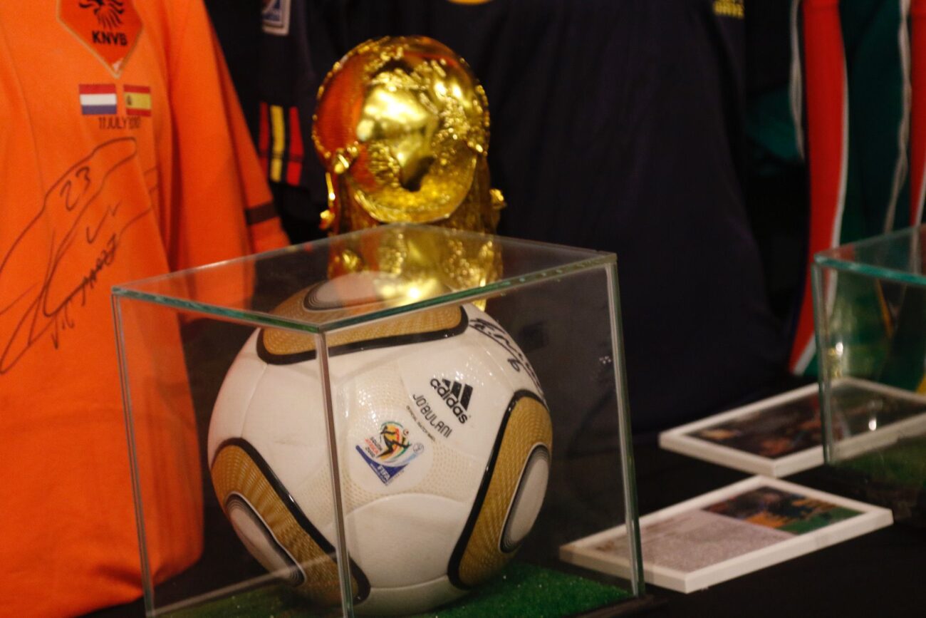 Soccer ball in a glass display case with a gold trophy resting on top, memorabilia in the background.